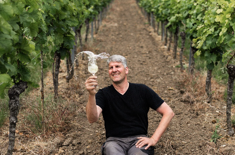 Achim Martin in den Weinreben mit einem Weißwein im Glas, das er kröftig schüttelt