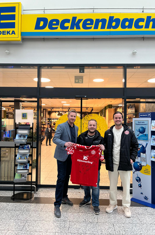 Marktinhaber Klaus Deckenbach (links) mit dem glücklichen Gewinner Herrn Presper und Herrn Laws (rechts) vom 1. FSV Mainz 05 bei der Übergabe des signierten Trikots vor dem Markteingang in Hartenberg